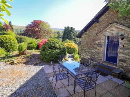 An outdoor patio with a glass table and metal chairs next to a stone building with a blue door at Stablemans Cottage at Stepping Stones in Ambleside