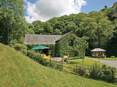 A house covered in ivy with a patio umbrella and a small gazebo in a grassy yard surrounded by trees at Riverside in Troutbeck