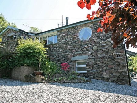 A stone exterior wall of a house with round and rectangular windows and gravel ground at Stone Howe in Ambleside