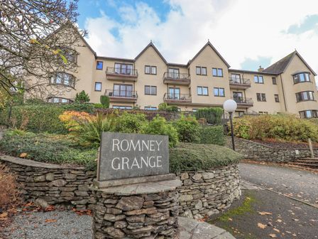 A multi story residential building with balconies and a stone sign saying Romney Grange surrounded by shrubs and trees at Waterhead Retreat Ambleside
