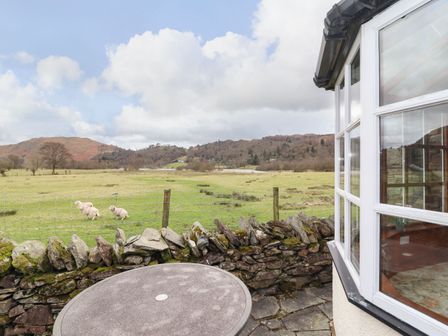 A stone patio with a round table next to a building window overlooking a fenced grassy field with sheep and hills at Pavement End Cottage in Grasmere