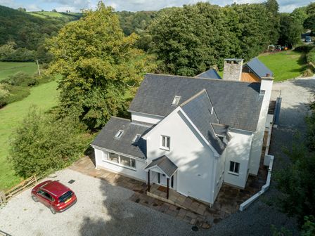 A white house with multiple gray slate roofs surrounded by trees and greenery with a red car parked on a gravel driveway at Priory House Barnstaple