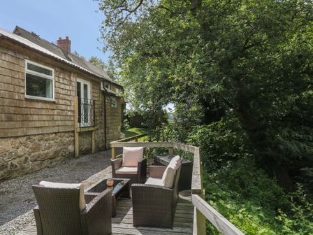 An outdoor wooden deck with wicker chairs and a table next to a house with wooden siding and surrounded by trees at Shilstone Lodge in Chagford