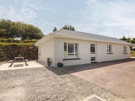 A single story white house with grey roof and windows and a wooden picnic table outside at Coolnaharragill Lower Annexe in Glenbeigh County Kerry