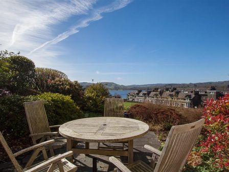 An outdoor patio with a round wooden table and chairs overlooking a row of houses and a lake at Romney 12 in Waterhead near Ambleside