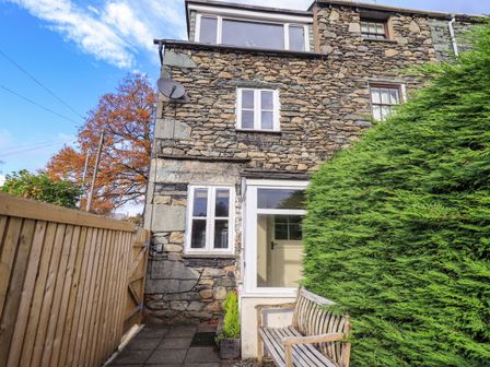 Stone exterior of a cottage with white windows a wooden bench a tall hedge and a wooden fence at Penny Red Cottage in Ambleside