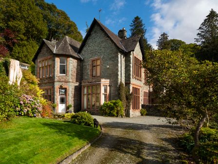 A stone house with large windows and a gravel driveway surrounded by trees and bushes at Brunt How in Ambleside