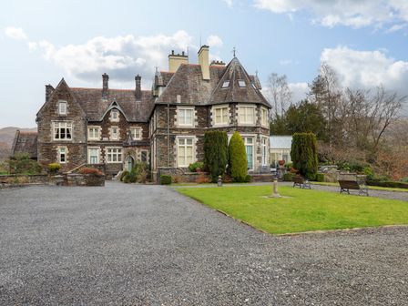 A large stone house with steep roofs and chimneys surrounded by gravel driveway and lawn with trees and benches