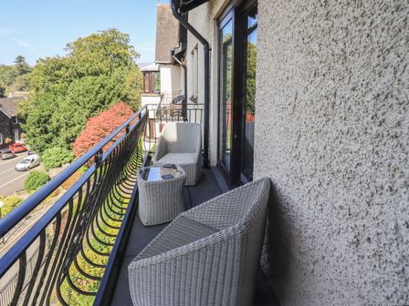 A balcony with wicker chairs and a small table overlooking trees and a street at Romney Grange in Ambleside