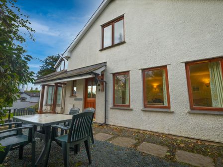 A patio area with plastic chairs and a table outside a white two-story house with wooden windows at Holly Cottage Ambleside