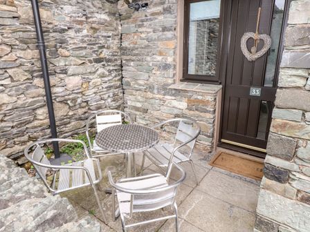 A small outdoor patio area with a round metal table and four metal chairs by a dark wooden door with the number 33 on a stone wall at Babbling Brook in Ambleside