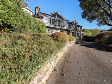 A stone house with a balcony and a driveway lined with bushes and trees at Garden House in Ambleside