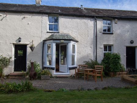 Exterior of a white textured wall cottage with black doors wooden chairs and table on gravel area and green plants at Swinside Cottage in Newlands Valley