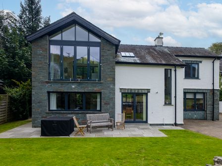 The back of a two-story house with stone and white walls a patio with wooden chairs and a bench and a green lawn at The Old Police House in Grasmere