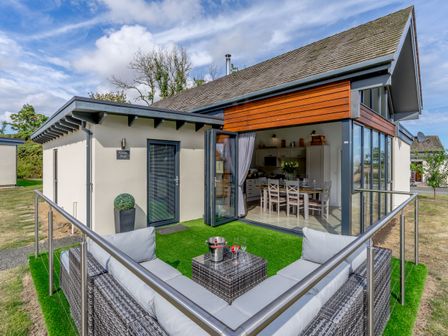 A small outdoor seating area with a sofa and table on artificial grass outside a house with open glass doors showing a dining table inside at Wood Sage in Bamburgh