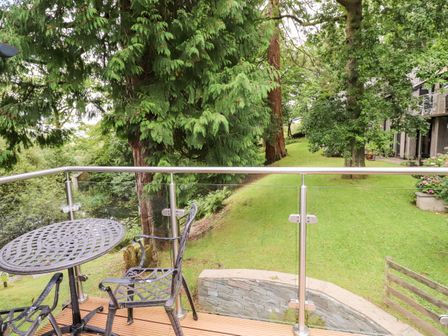 An outdoor seating area with a metal table and two chairs on a wooden deck overlooking a grassy area with trees at Fir Howe in Skelwith