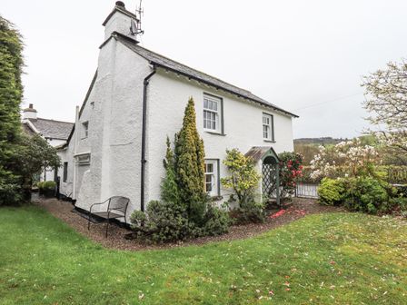 A white cottage with green-trimmed windows surrounded by garden with various shrubs and a lawn at Syke Cottage in Hawkshead