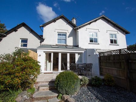 The back of a two story white house with patio doors leading to a small patio with a metal table and four chairs surrounded by bushes and a stone path at Mylnbeck in Bowness