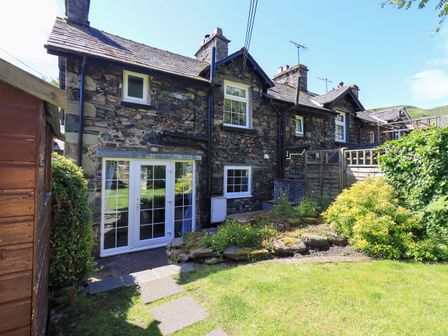 The back exterior of a stone cottage with white framed windows and doors and a garden with grass and shrubs at Bleaberry Cottage in Glenridding