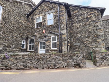 A stone cottage with a white door and three windows surrounded by a low stone wall at Gildabrook Cottage in Bowness