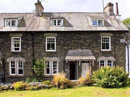 A stone house with multiple windows and a small porch surrounded by grass and bushes at Rowan Cottage in Hawkshead