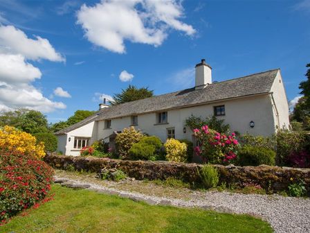 A white cottage with chimneys surrounded by flowering bushes and a stone wall at Old Droomer in Windermere