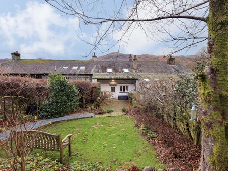 A garden with a wooden bench and hedges in front of a row of cottages with skylights and chimneys at Daisy Cottage in Grasmere