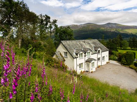 A white two-story house with slate roof surrounded by trees and purple flowers with mountains in the background at Kirkstones in Keswick