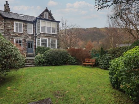 A garden with green lawn bushes and a wooden bench outside a stone house at Fairy Glen in Grasmere near Ambleside