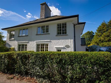 A two-story white house with multiple windows behind a trimmed hedge under a blue sky at Crozier Cottage in Grasmere
