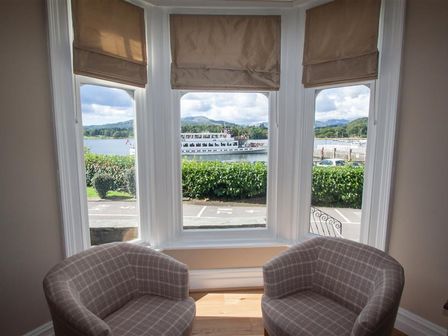 Two plaid armchairs facing a bay window with beige roman blinds overlooking a road hedge and a boat on water at Waterside Ambleside