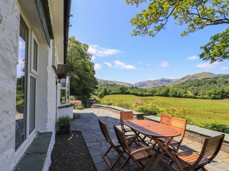 An outdoor patio with wooden table and chairs overlooking a grassy field and hills at Spindle Coppice in Skelwith Bridge