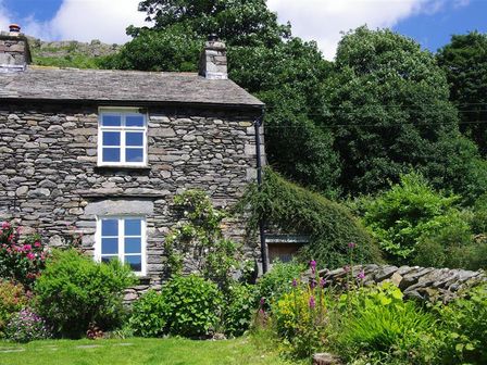 A stone cottage with white framed windows surrounded by green bushes and flowers in a garden at Thomas Cottage in Hartsop
