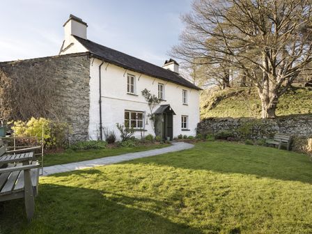 A white stone house with a slate roof and chimneys surrounded by a garden with benches and a large tree at Yew Tree Farm in Coniston