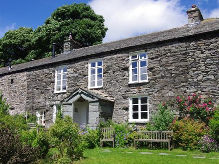 A stone house with white framed windows and a porch surrounded by green plants and wooden benches at Thomas Grove House in Ullswater