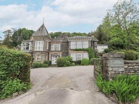 A stone house with multiple windows and a pointed turret surrounded by greenery with a gravel driveway and a signpost at The Writing Room in Hawkshead