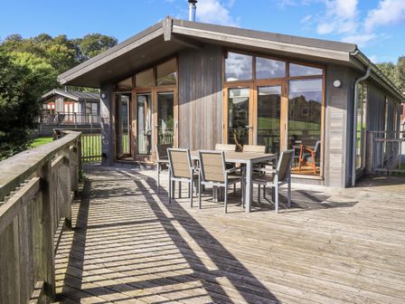 A wooden deck with a table and six chairs outside a cabin with large windows at How Beck in Hawkshead