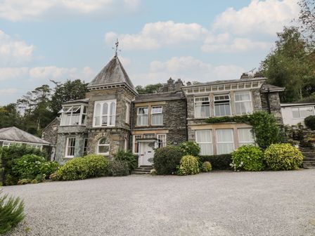 A stone building with multiple windows and a tower surrounded by bushes and gravel in front at The Rockery Suite in Hawkshead