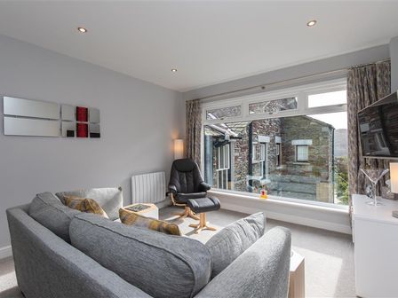 A living room with a gray sofa, a black chair, a small table, wall art, and a large window showing a stone building at Silver Howe in Grasmere
