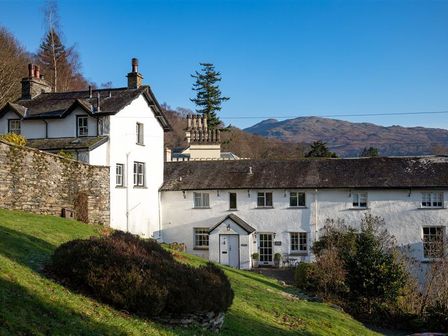 A white stone house with multiple chimneys on a grassy hill with mountains in the background at The Hayloft in Ambleside