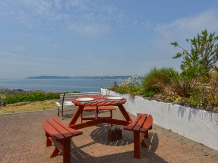 An outdoor patio with a round wooden picnic table set with plates and glasses overlooking the sea at SoundView in Down Thomas