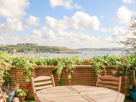 An outdoor patio with wooden table and chairs overlooking a body of water with boats and green hills at Through The Porthole in Falmouth