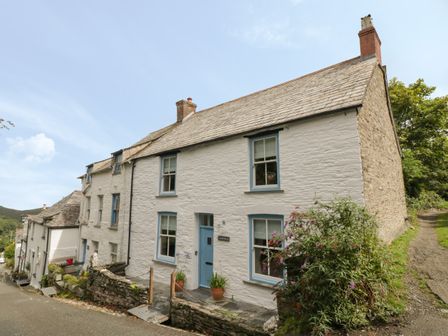 A white stone house with blue window frames and a blue door on a sloped street at Fairfield Cottage in Boscastle