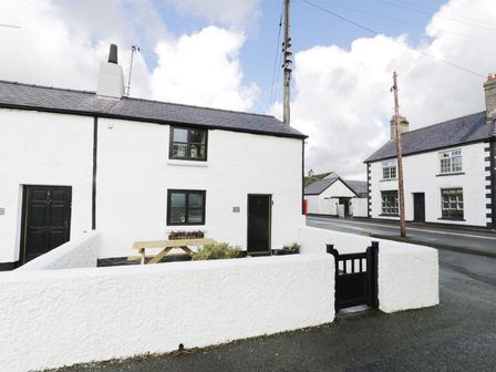 A white cottage with black doors and windows a wooden picnic bench and a gated front yard at Menai Cottage in Brynsiencyn