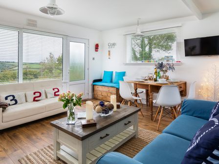 A living room with a white sofa and blue cushions a wooden coffee table and a dining table with white chairs and tableware at The Hut in Dartmouth
