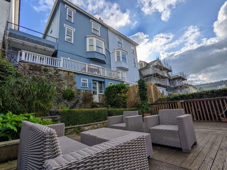 An outdoor patio with wicker furniture and a blue multi-story house with a stone base in the background at Armorel House in Dartmouth