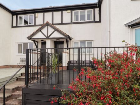 A white two story house with black trim and a covered porch with two black doors a black metal railing around a deck and red flowering bushes in front at Castle View in Deganwy