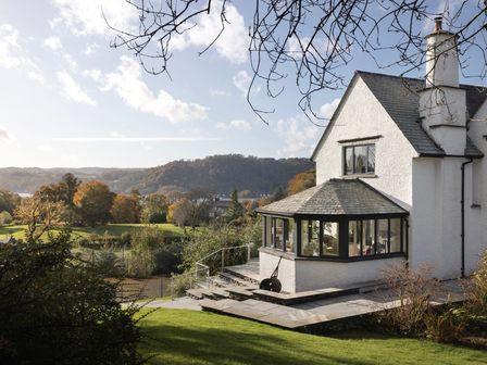 A white house with a slate roof and large windows overlooking a green landscape with trees and hills at Cherry Trees in Bowness