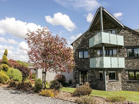 A stone house with glass balconies surrounded by trees and bushes at The Golf Apartment in Kendal