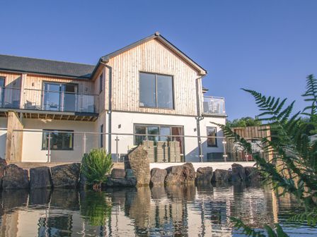 An exterior view of a two story house with wooden and white walls glass balcony railings and a pond with rocks and plants in front at Swallows Dip in Honiton